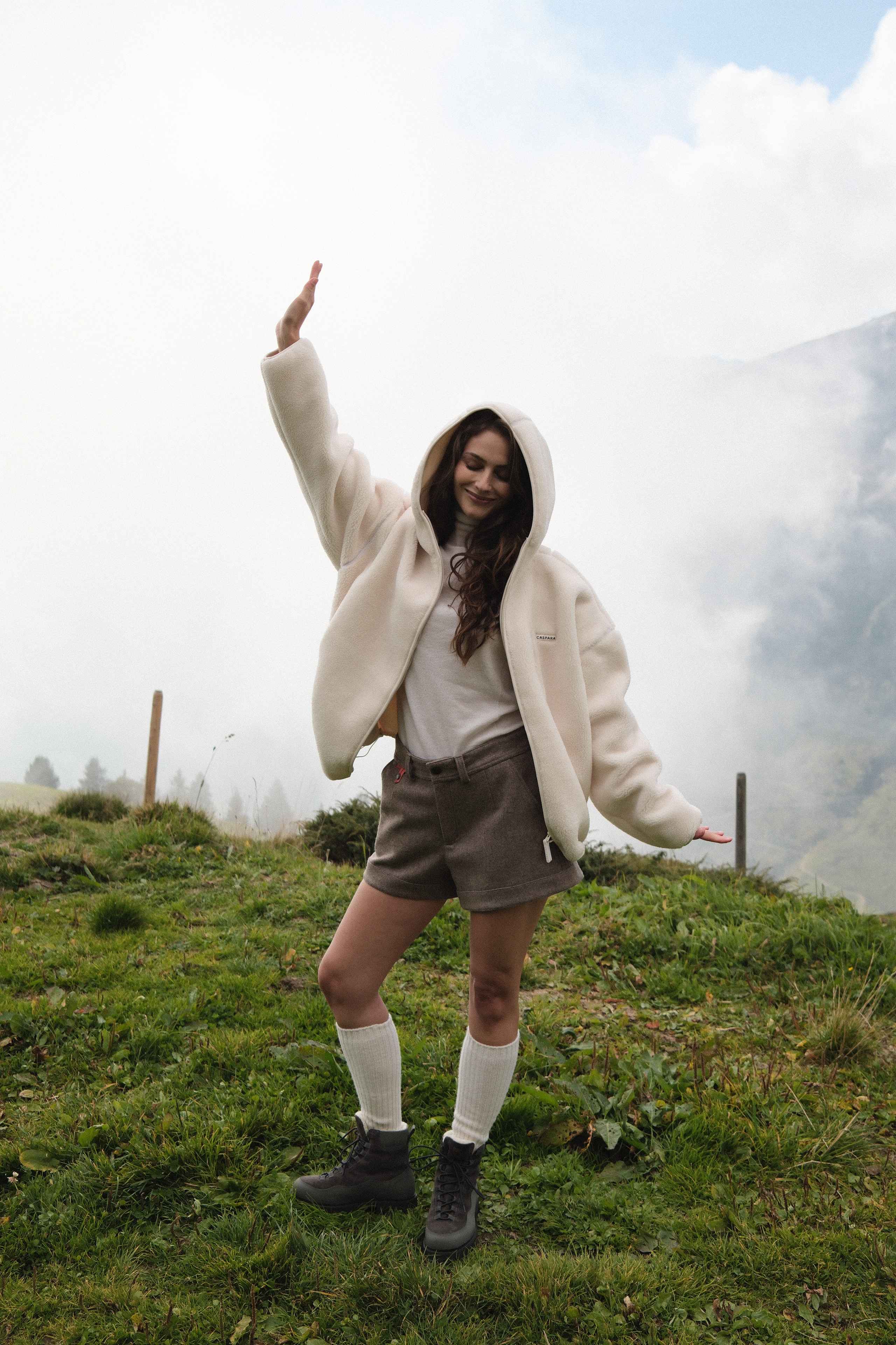 Woman wearing a creme CASPARA Cloudfleece hooded jacket outdoors on a misty hillside.